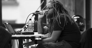 D' de Kabal - Felures D' de Kabal assis à un bureau parlant dans un micro, photo en noir et blanc